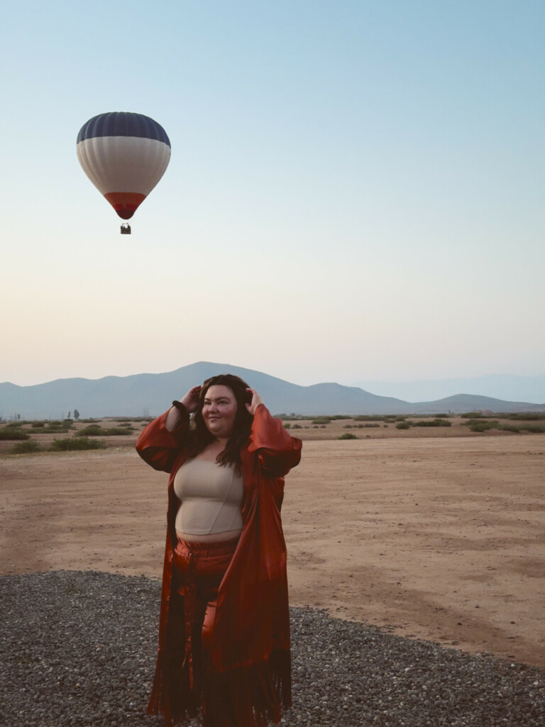 Plus size woman wearing a matching satin pant set with a kimono in the desert of Morocco while a hot air balloon takes off in the distance